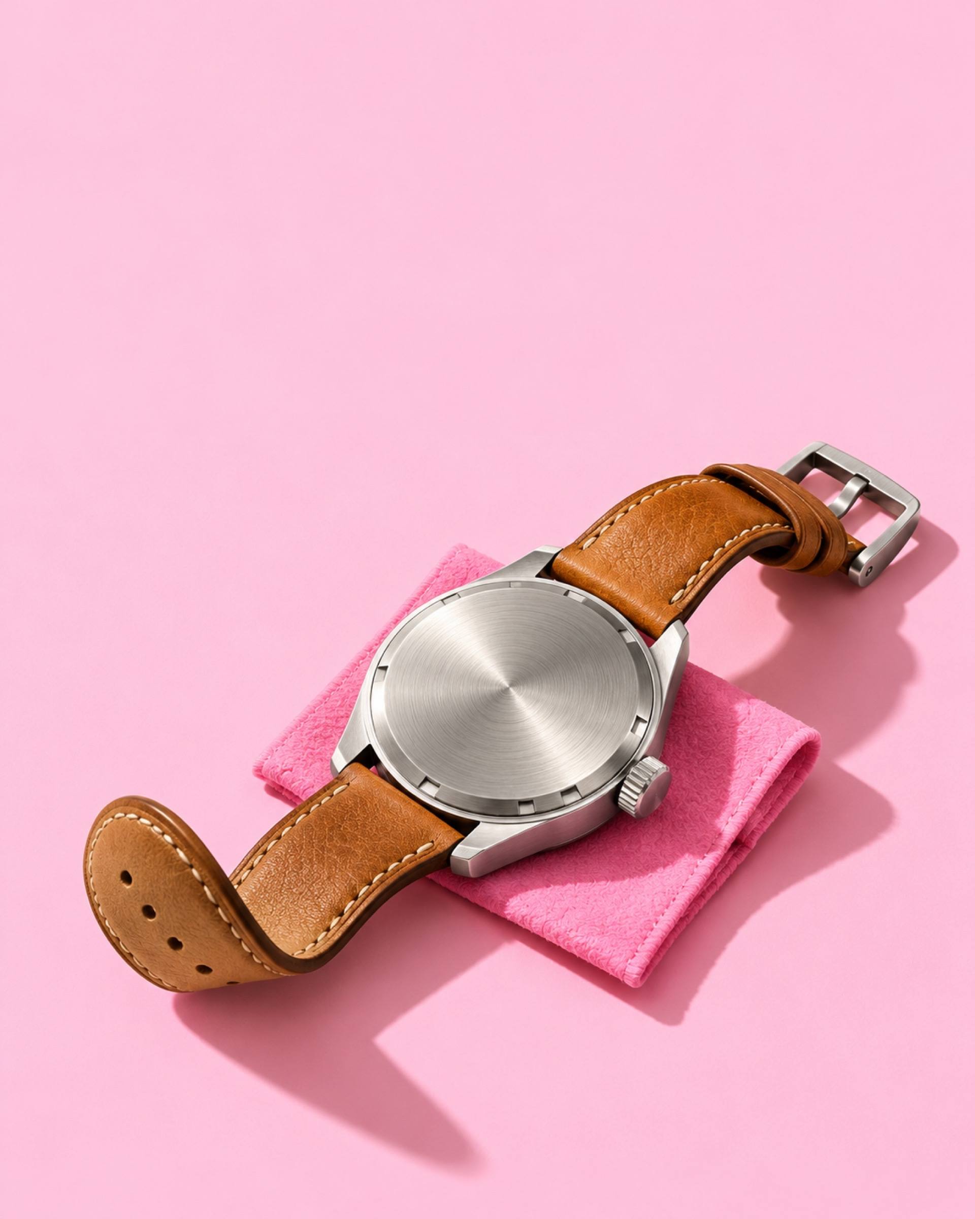 The back of a steel watch on a tan leather strap, resting on a pink cloth